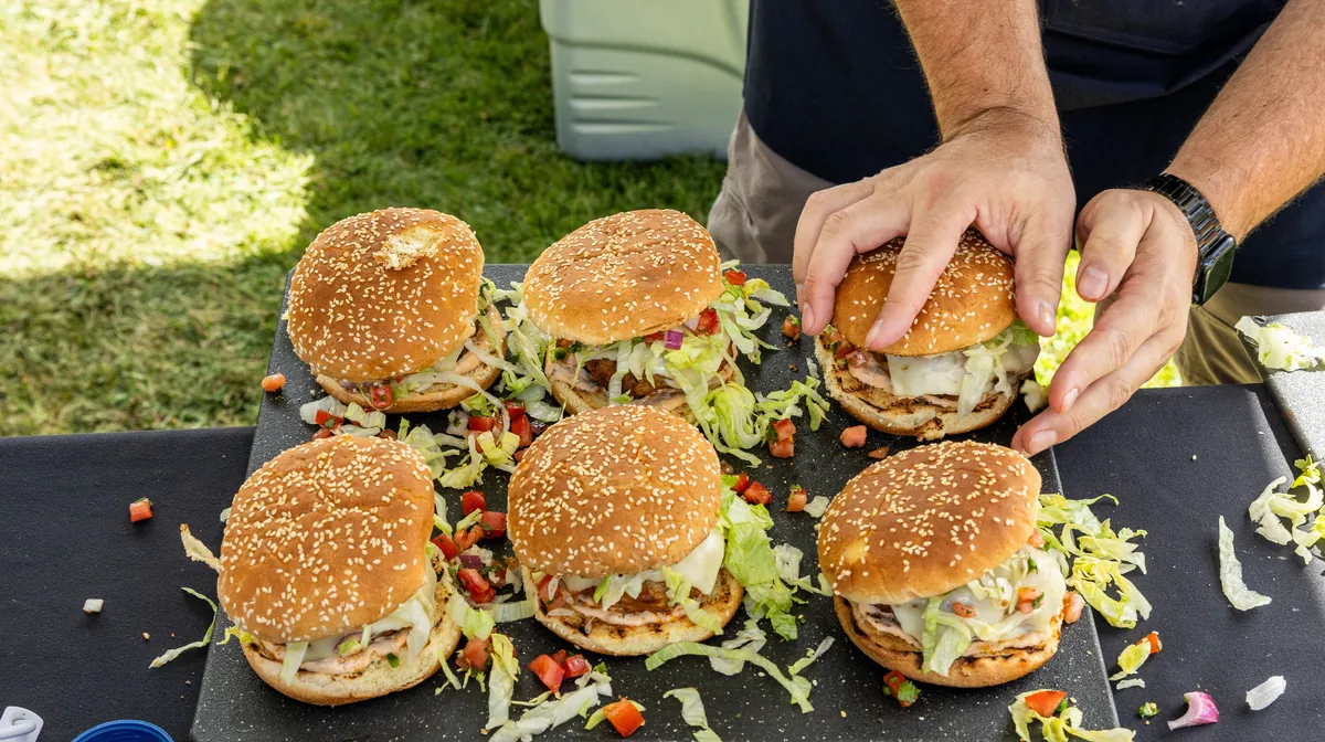 Clark Neal assembling Smokey Firebird Burgers on sesame seed buns with shredded lettuce and diced tomato on a black serving board